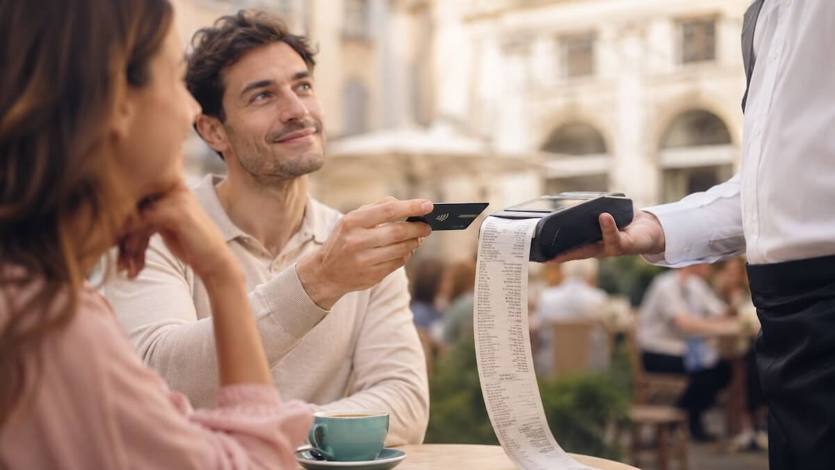A close-up of a couple at a quaint European café; one person is confidently holding out a credit card to a waiter holding a payment terminal, while the other partner relaxes in the foreground looking completely unbothered. A subtle teal (#00A59B) ceramic coffee cup sits on the table.