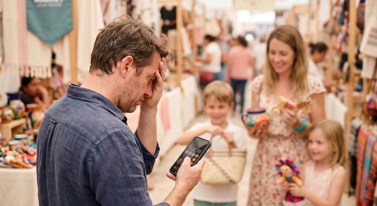 An over-the-shoulder shot of an exhausted dad holding his head in slight frustration while looking at a calculator app on his phone, surrounded by his family holding up souvenirs in a vibrant local market.